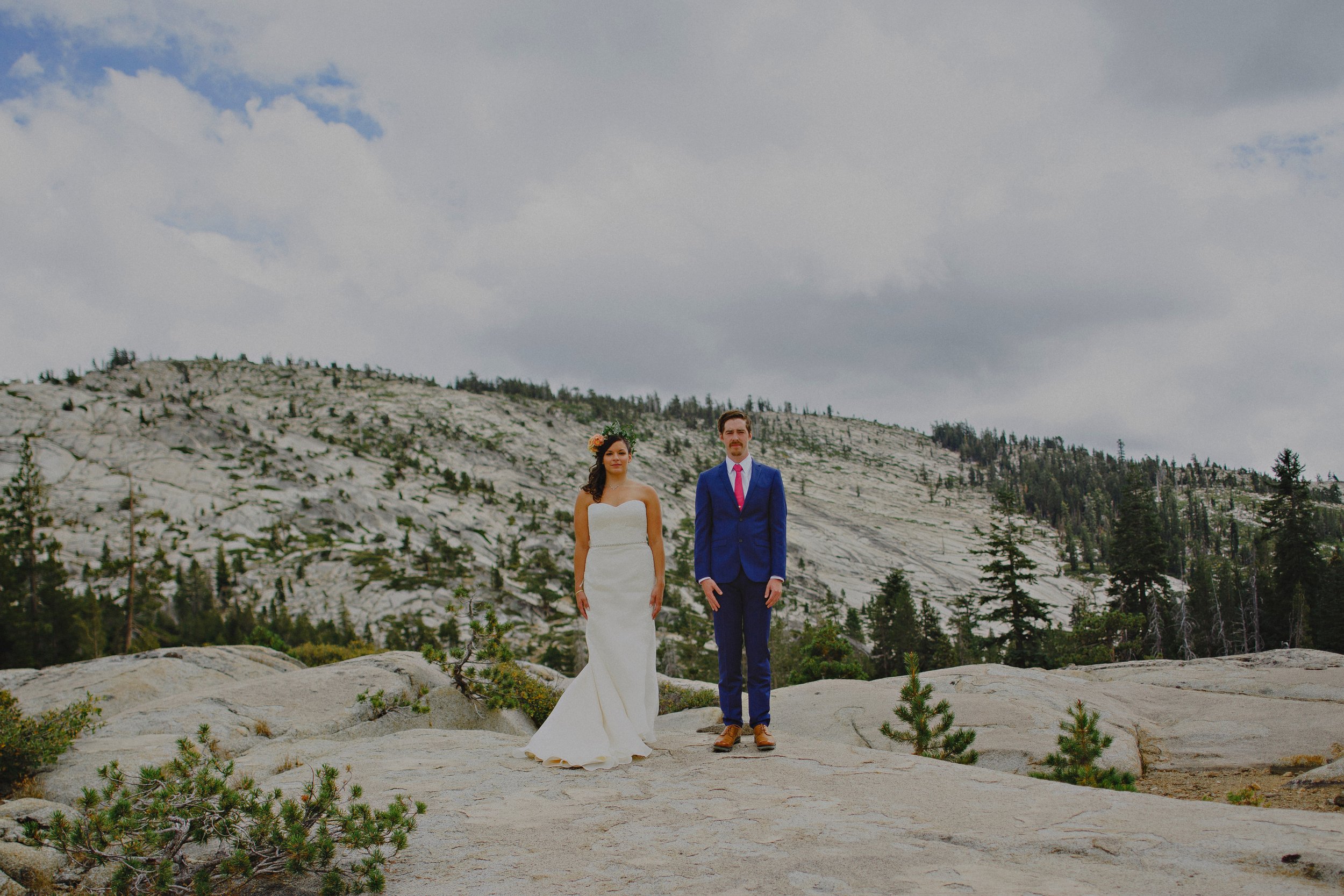 A bride in a white dress and a groom in a blue suit with a red tie stand on a rocky terrain with a mountainous background and cloudy sky.