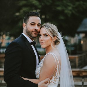 A bride in a white dress and veil and a groom in a black tuxedo stand closely together outdoors.