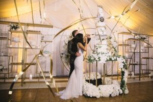 A couple in wedding attire cuts a large tiered cake adorned with flowers and a clock ornament, surrounded by gold ribbon decorations in an elegant indoor setting.