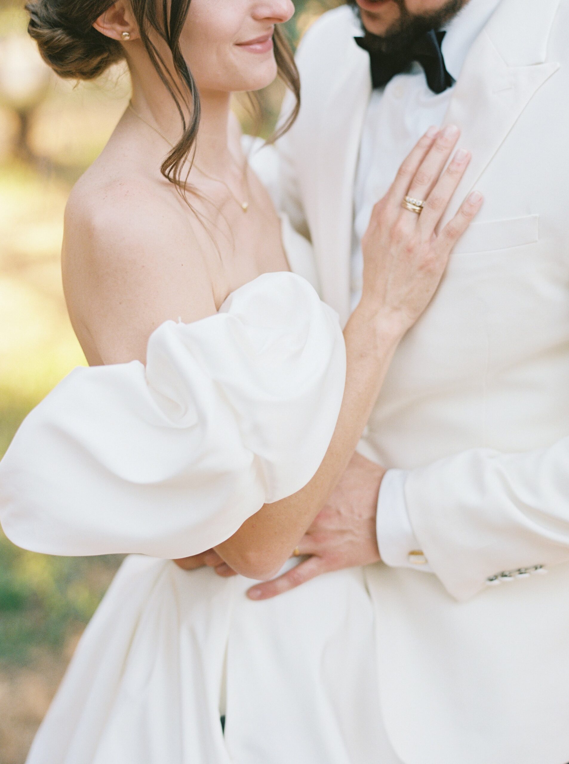 A couple stands close together, wearing matching white formal attire. The woman has off-the-shoulder puff sleeves, and the man wears a bow tie. They are smiling, with her hand resting on his chest.