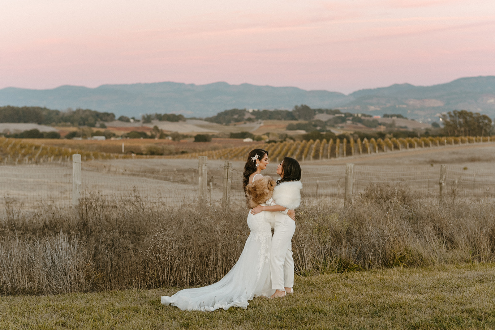 Two women in formal attire embrace in a field during sunset, epitomizing elegance. One wears a long white dress, the other dons white pants with a fur stole—an exquisite scene set against Napa Valley's rolling hills, reminiscent of a wedding planner's dream come true.