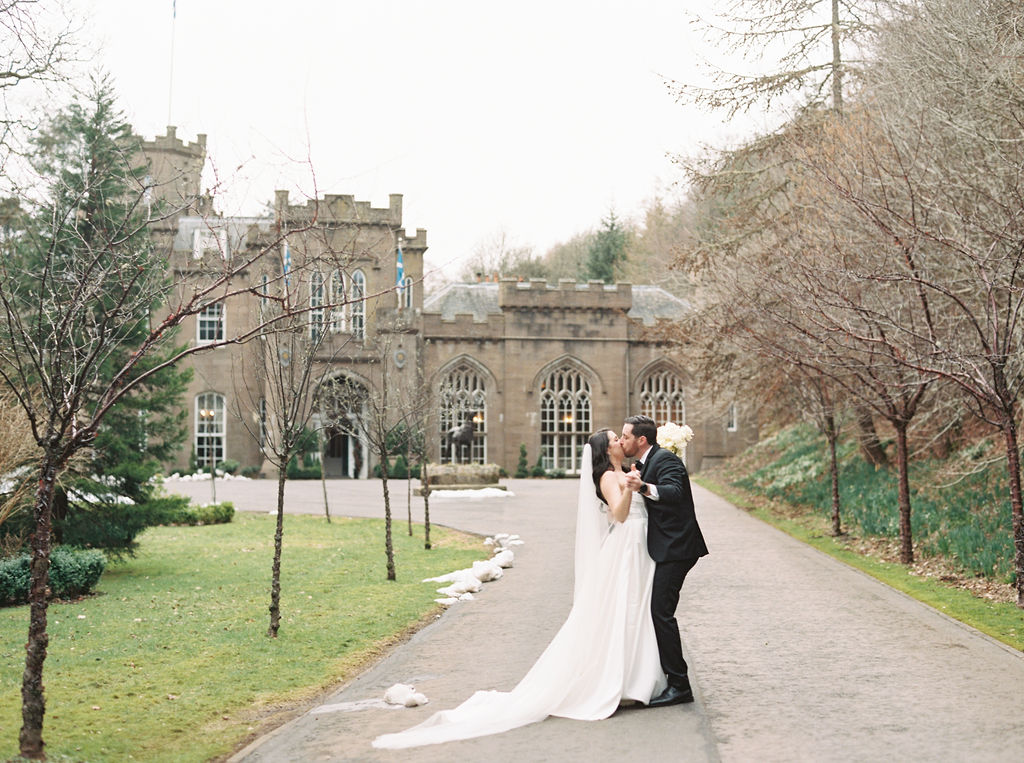 A couple in wedding attire shares a kiss on a path with a historic castle in the background, expertly orchestrated by destination wedding planners. Bare trees line the walkway, and the surroundings are lush with greenery.