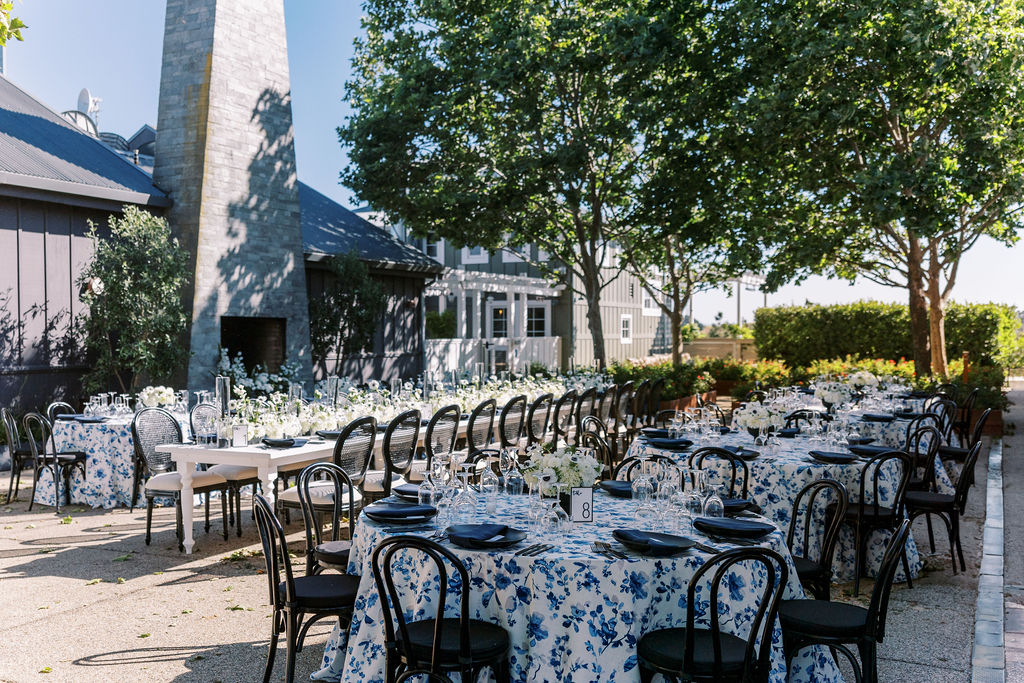 Outdoor dining setup for a Napa wedding, featuring round tables adorned with white and blue floral tablecloths, black chairs, and charming floral centerpieces. The tables are elegantly arranged near a building amid lush greenery and trees.
