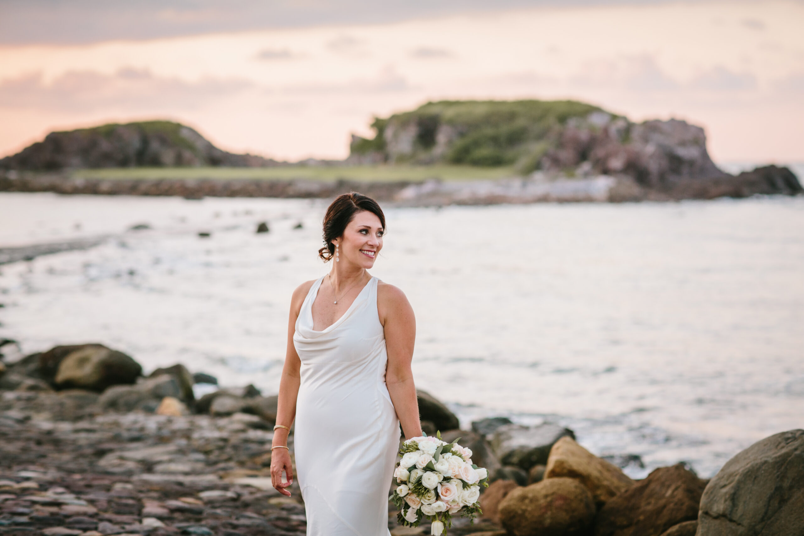 A woman in a white dress stands on a rocky beach holding a bouquet, with an ocean backdrop and small islands in the distance at sunset.