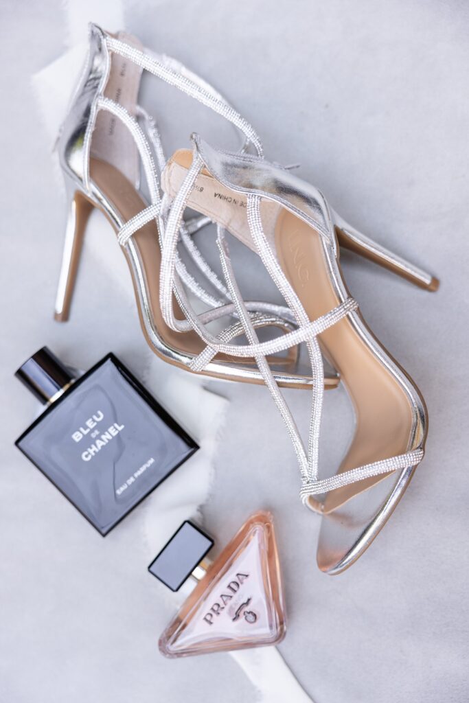 Silver high-heeled sandals beside bottles of Bleu de Chanel and Prada perfume on a light surface.