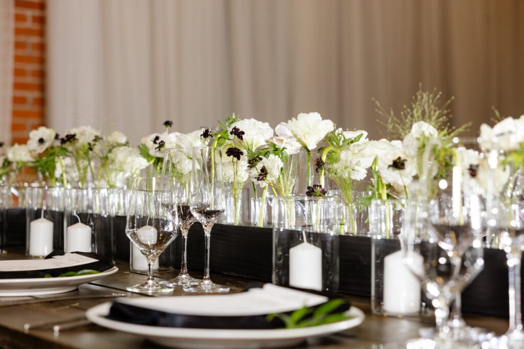 Table set with plates, wine glasses, white candles, and a centerpiece of white and dark flowers in clear vases, against a backdrop of white curtains and a brick wall.