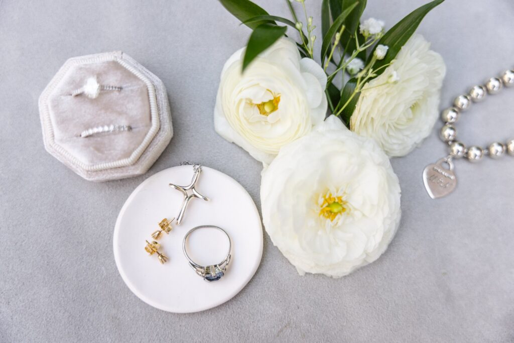 A white velvet ring box, gold and silver jewelry on a dish, white flowers, and a silver bead bracelet with a heart charm arranged on a light gray surface.