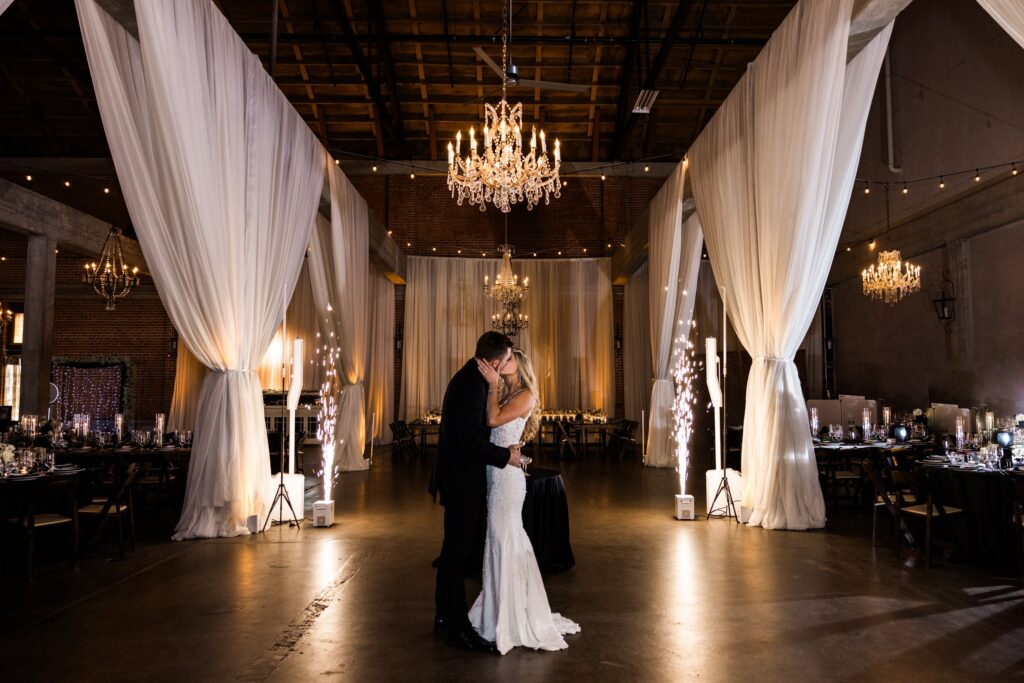 A bride and groom share a dance in an elegant, dimly lit venue with chandeliers, draped white fabric, and decorated tables.
