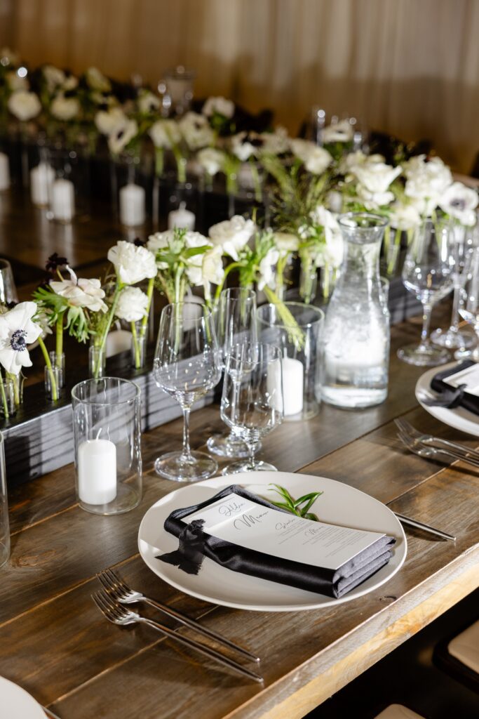 Elegant table setting with white plates, glasses, black napkins, candles, a water carafe, and white floral centerpieces on a wooden table.