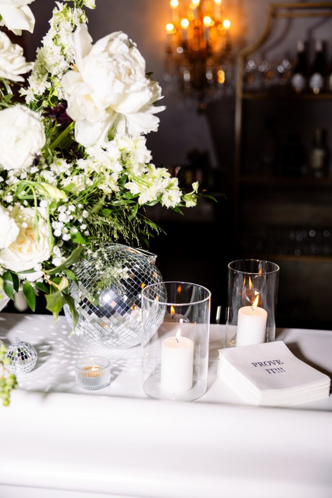 White flowers, a disco ball, lit candles in glass holders, and a stack of napkins with “PROVE IT!” printed on them arranged on a white surface.