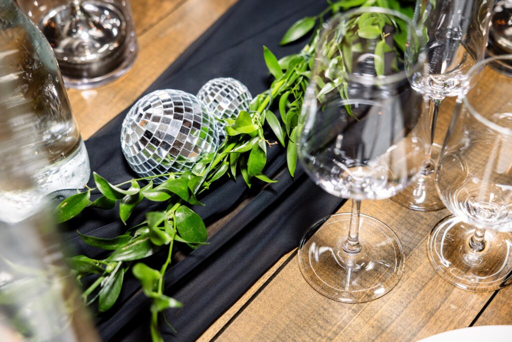 Table setting with wine glasses, a water bottle, green foliage, and small mirrored disco balls on a black cloth runner over a wooden surface.