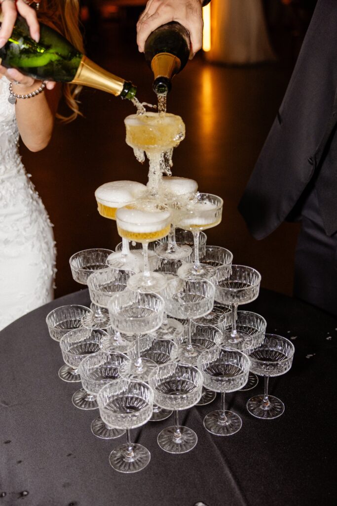 Two people pour champagne into the top glasses of a pyramid-shaped tower of coupe glasses, causing the champagne to overflow into the glasses below.