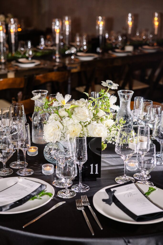 Round table set for an event with white flower centerpiece, glassware, candles, menus, and black tablecloth. Table number 11 is displayed in the center.
