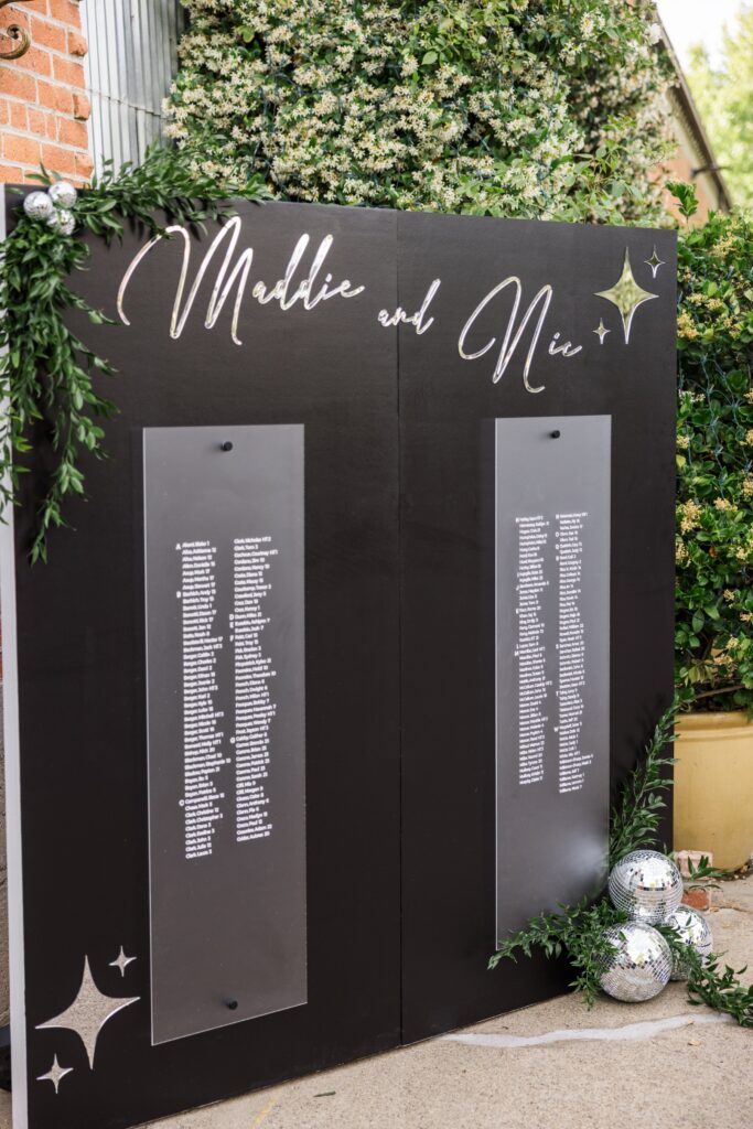 Black wedding seating chart display with “Maddie and Nic” written at the top, decorated with greenery and silver disco balls, positioned outdoors near a brick wall and plants.
