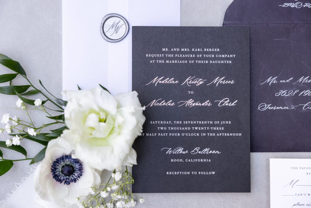 A black wedding invitation card with white script text lies on a light surface, decorated with white flowers and greenery, alongside matching envelopes and a wax seal.