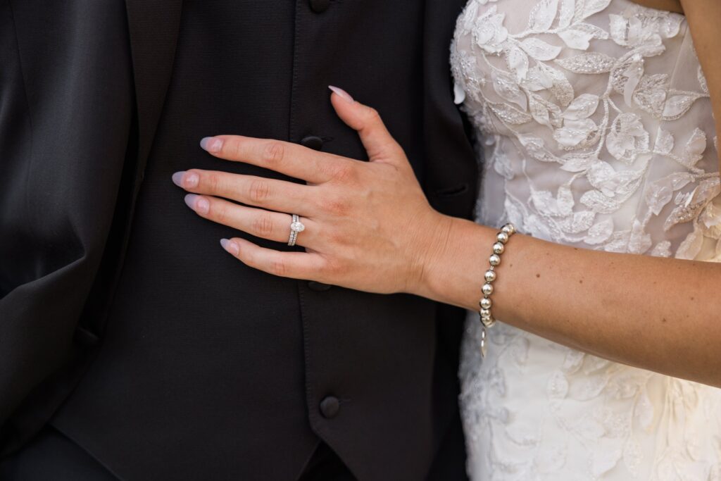 A close-up of a bride’s hand with a wedding ring resting on a groom’s black suit. The bride wears a white lace dress and a pearl bracelet.