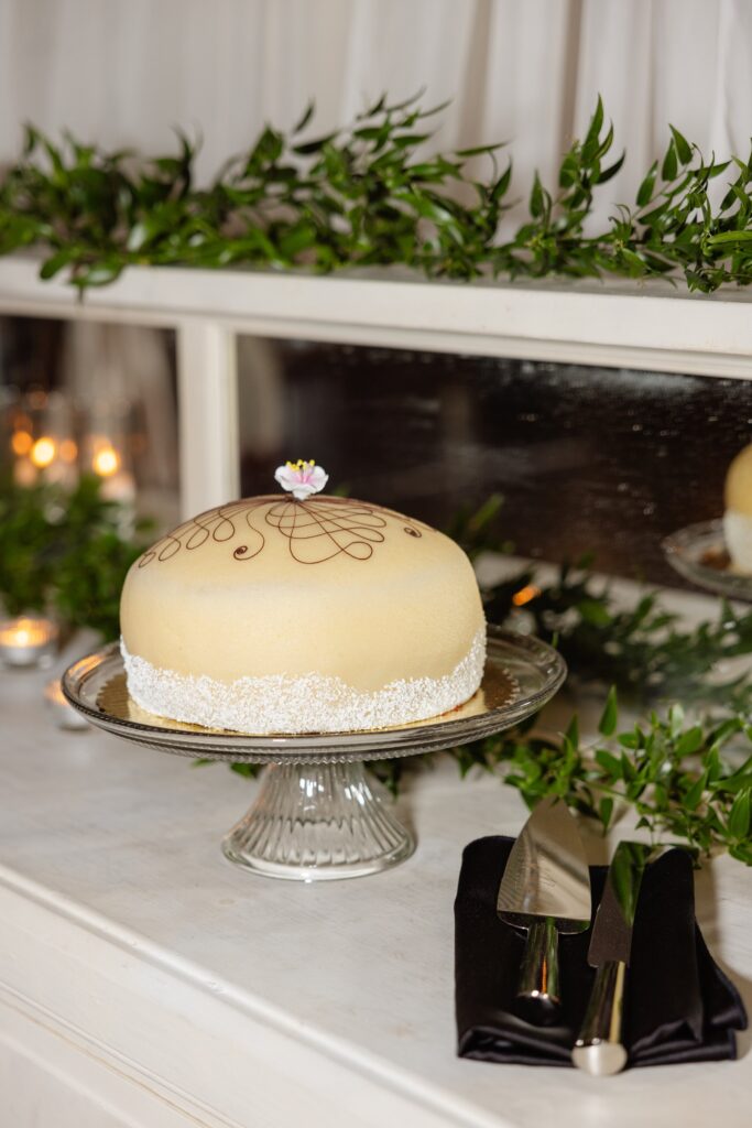 Round cake with white frosting and decorative swirl, topped with a small flower, on a glass stand beside a cake knife and greenery on a white table.