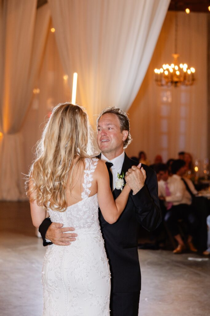 A man in a black suit dances with a woman in a white lace dress in a decorated ballroom with chandeliers and draped curtains.