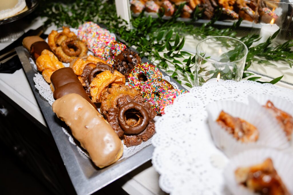 A tray of assorted donuts and pastries, some with chocolate and rainbow sprinkles, displayed on a decorated table with greens and a lace doily.
