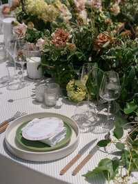 A table set with plates, cutlery, glasses, candles, and a napkin, surrounded by lush green and brown floral arrangements.