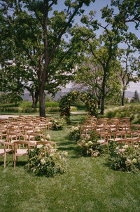 Outdoor wedding ceremony setup with rows of wooden chairs, floral arrangements, and a flower-decorated arch under tall trees on a grassy lawn.
