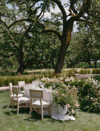 A table set for four with white chairs and floral arrangements sits on grass under large trees in an outdoor garden setting.
