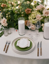 Elegant table setting with plate, green charger, cutlery, glasses, candles, and floral centerpiece featuring roses and greenery on a white tablecloth.