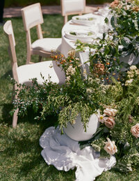 Outdoor table with white chairs, white tablecloth, place settings, and a large floral arrangement with greenery and light-colored flowers on green grass.