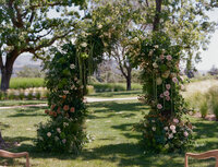 A floral wedding arch covered in greenery and roses stands on a grassy lawn, with trees and fields in the background.