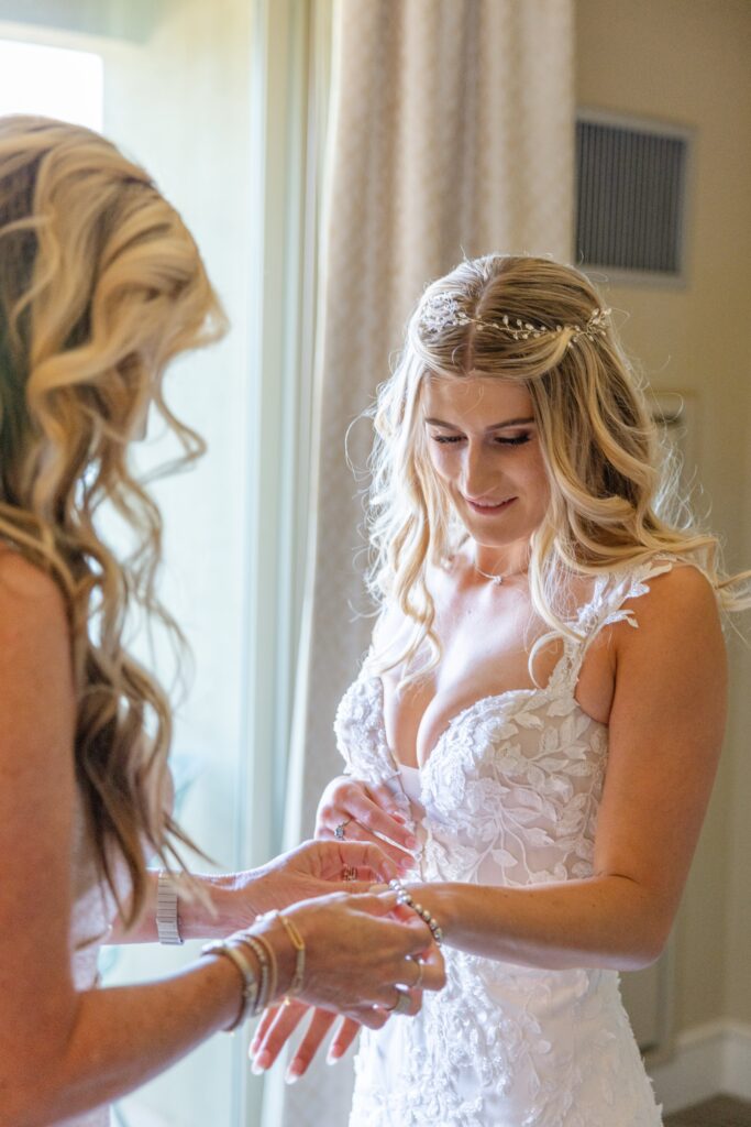 A woman in a white lace wedding dress stands indoors while another woman helps fasten a bracelet on her wrist.
