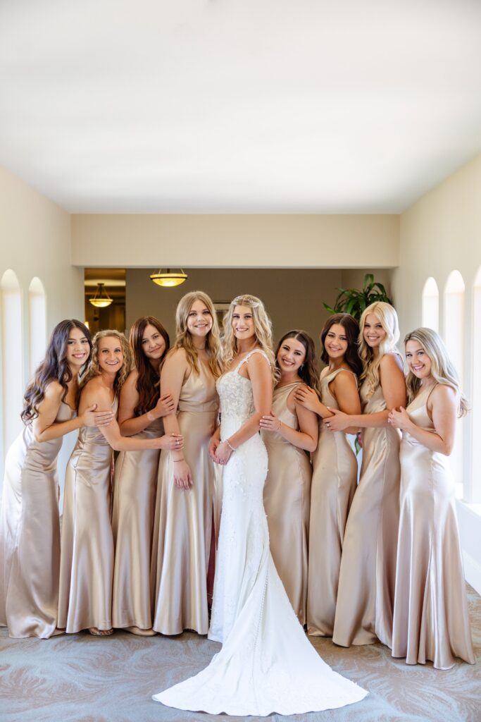 A bride in a white dress stands with seven bridesmaids in matching champagne gowns, all smiling and posing indoors in a bright hallway.