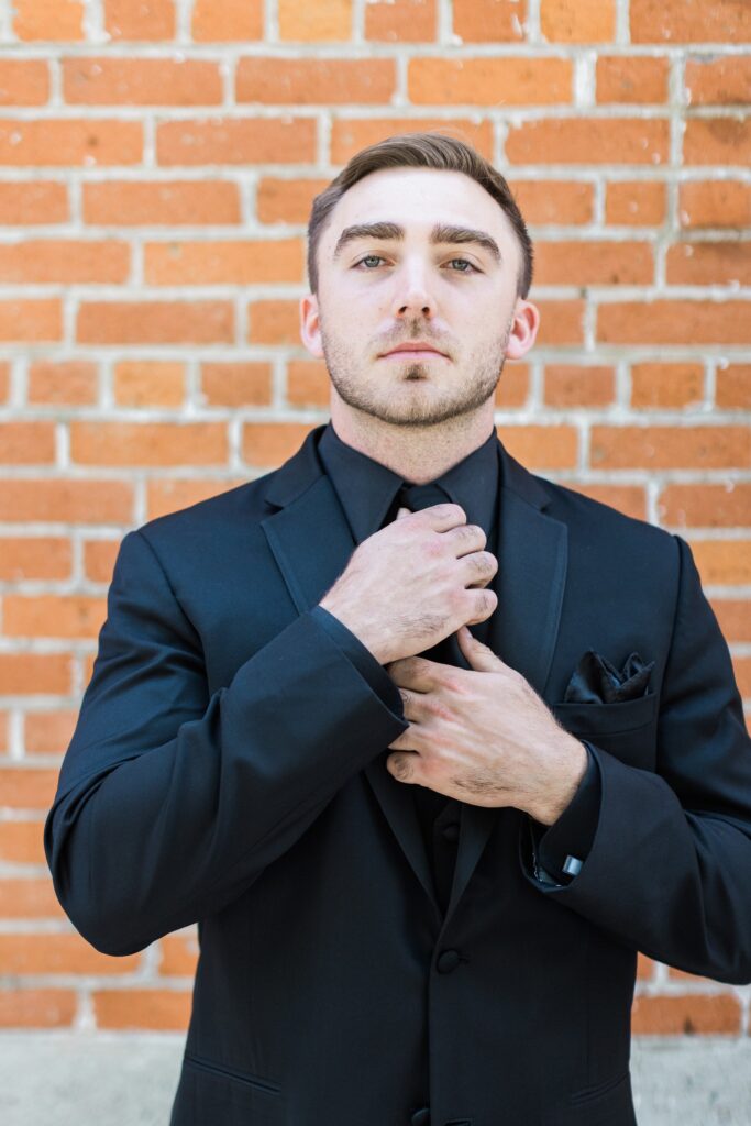A man in a black suit adjusts his tie while standing in front of a red brick wall.
