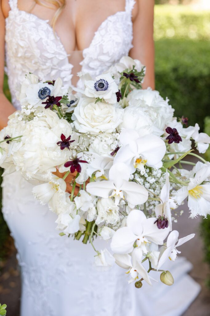 Bride in a white dress holding a bouquet of white orchids, roses, and anemones with small dark purple flowers.