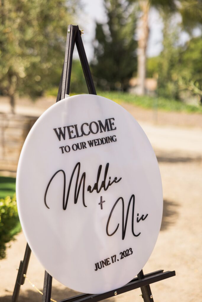 Round wedding welcome sign on an easel outdoors, reading “Welcome to our wedding, Maddie + Nic, June 17, 2023” in black script and block letters.