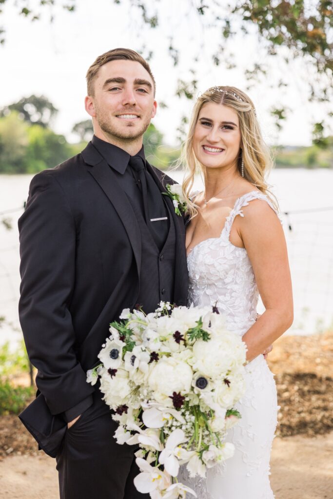 Bride in a white dress holding a bouquet stands next to groom in a black suit, posing outdoors by a body of water.