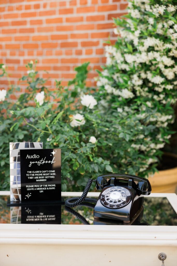 A black rotary phone sits on a table outdoors next to a sign with instructions, with green plants and a brick wall in the background.