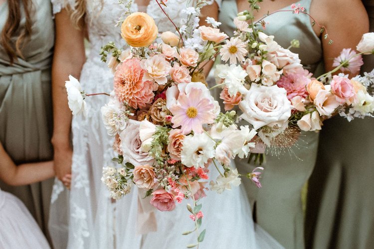 A bride holding a large, colorful bouquet of flowers stands with bridesmaids in green dresses.