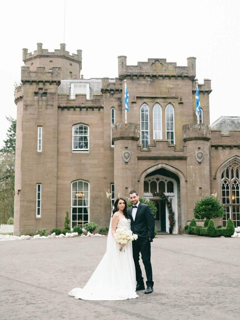 A bride in a white dress and a groom in a black suit stand in front of a large castle-like building holding a bouquet of white flowers.