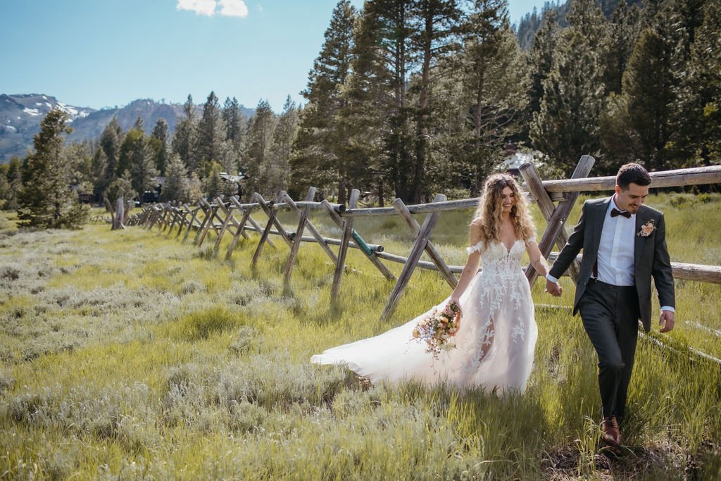 A bride and groom walk hand in hand in a grassy field near a wooden fence. The bride holds a bouquet and wears a white dress, while the groom is in a dark suit with a bow tie.
.