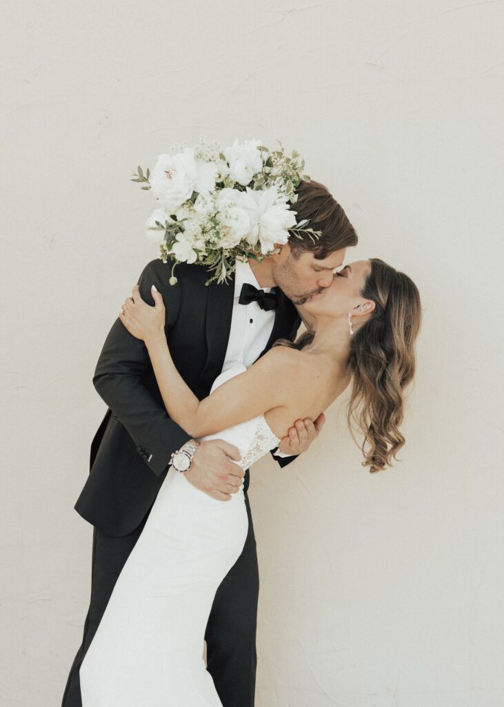 A couple dressed in formal wedding attire share a kiss, with the groom dipping the bride backward. The bride holds a bouquet of white flowers.