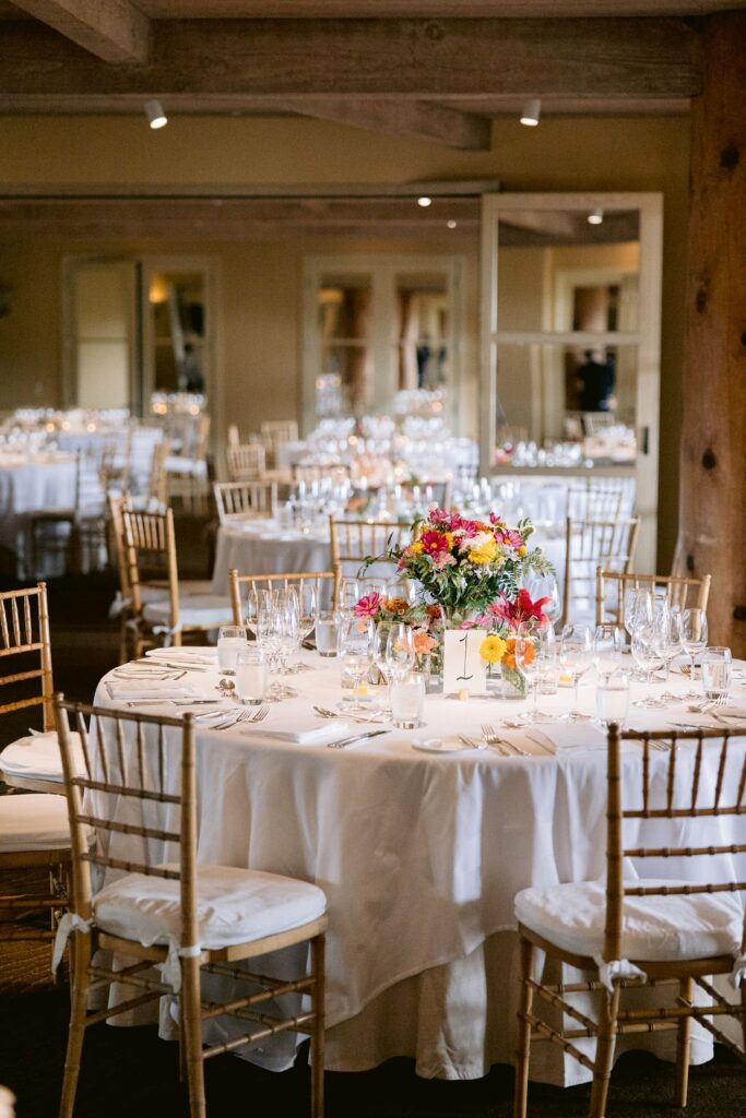 A round table set for a formal event with white tablecloth, gold chiavari chairs, silverware, glassware, and a floral centerpiece.