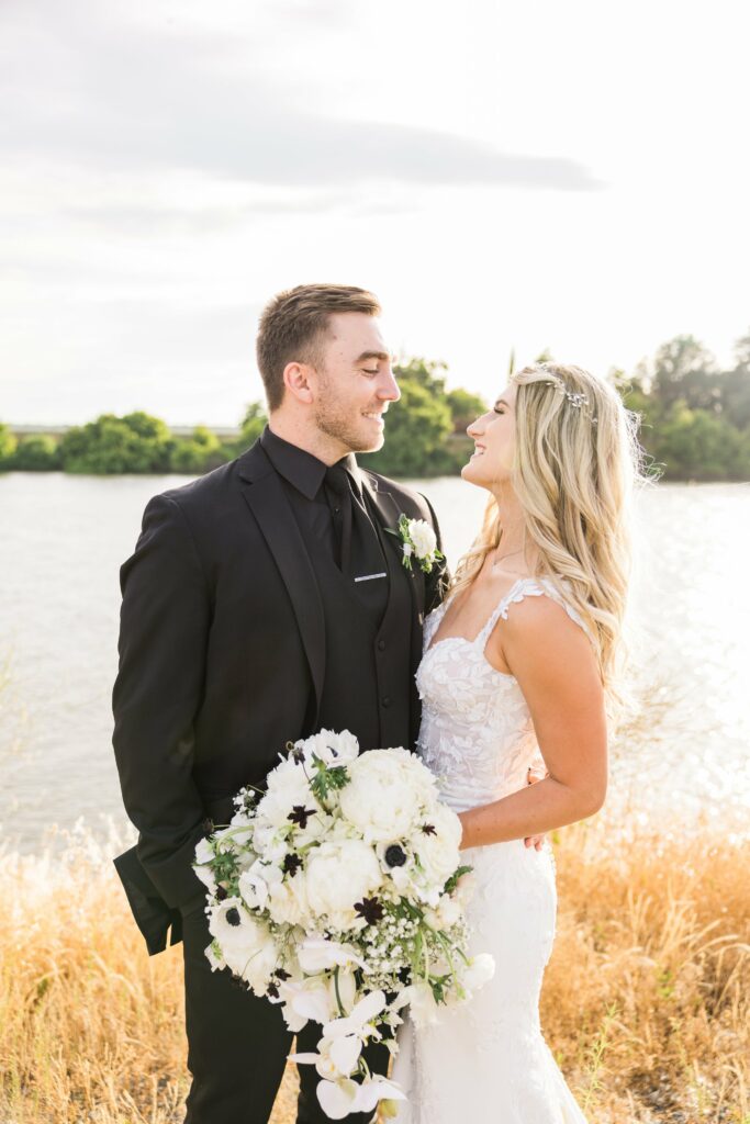 A bride and groom stand together outdoors by a body of water. The groom is wearing a black suit and the bride is in a white dress holding a bouquet of white flowers. They are gazing at each other and smiling.