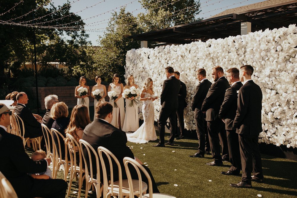 A wedding ceremony outdoors on a sunny day with a bridal party and groomsmen standing near a floral backdrop. Guests are seated on wooden chairs watching the couple.