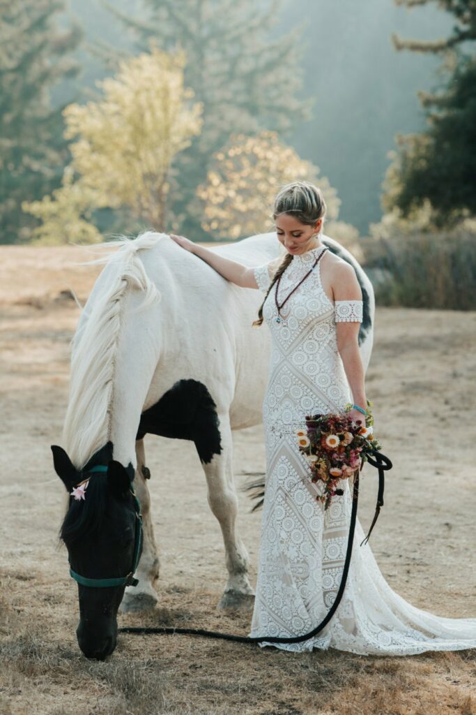 A bride in a lace gown holds a bouquet and stands beside a grazing white horse in a scenic outdoor setting.
