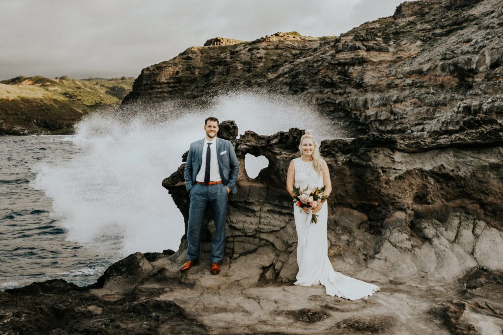 A man in a gray suit and a woman in a white dress stand on a rocky shoreline with crashing waves in the background. The woman holds a bouquet of flowers.