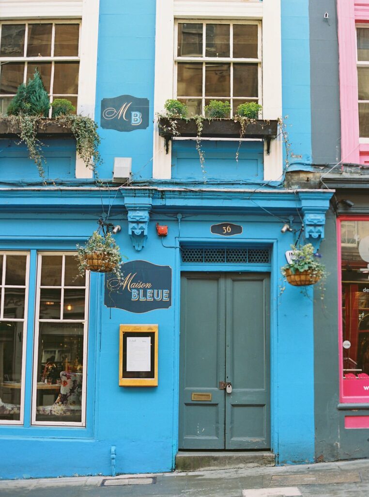 A brightly painted blue storefront, named Maison Bleue, with a black door, hanging plants, and window planters. The adjacent building on the right is pink. The number 36 is above the door.