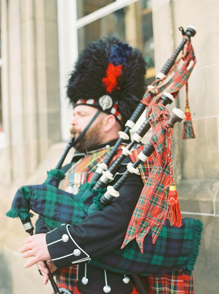 A bearded person dressed in traditional Scottish attire plays the bagpipes along a stone building.