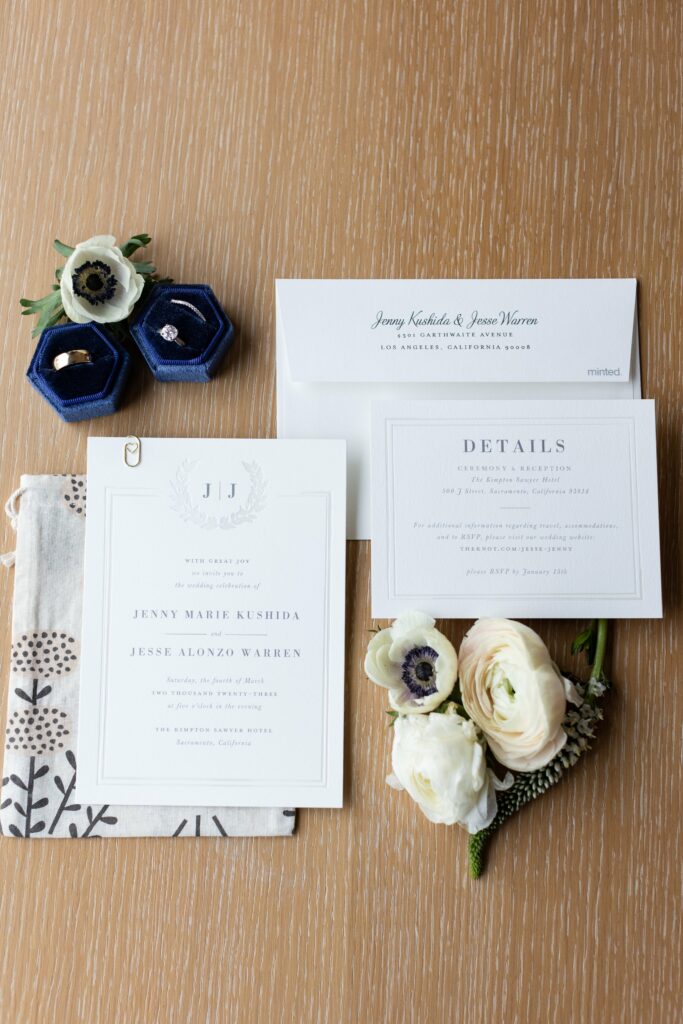 Wedding invitation set with floral accents laid out on a wooden surface. Items include the main invite, detail card, RSVP envelope, decorative fabric, rings in blue boxes, and white flowers.