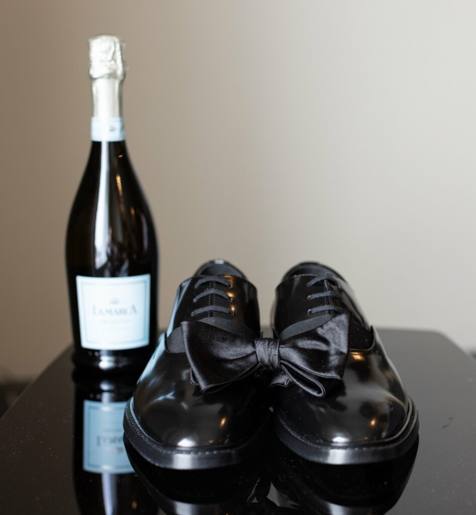 A pair of shiny black dress shoes with a black bow tie and a bottle of sparkling wine on a reflective surface.