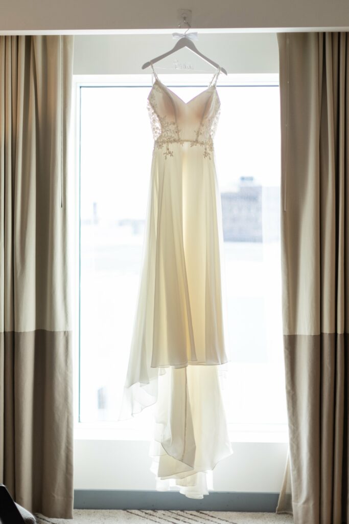 A white wedding dress with delicate straps and lace details hangs on a hanger in front of a window with beige curtains.